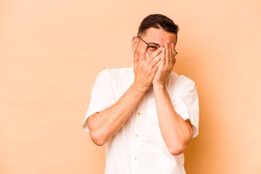 Latin kökenli genç adam bej arka planda izole edilmiş. Kameraya parmaklarıyla göz kırpıyor. Yüzünü kapatmaktan utanıyor..