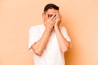 Latin kökenli genç adam bej arka planda izole edilmiş. Korkmuş ve gergin parmaklarıyla göz kırp..