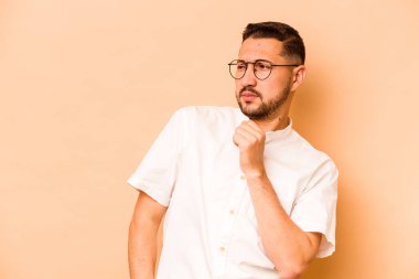 Latin kökenli genç adam bej arka planda izole edilmiş kafası karışmış, şüpheli ve güvensiz hissediyor..