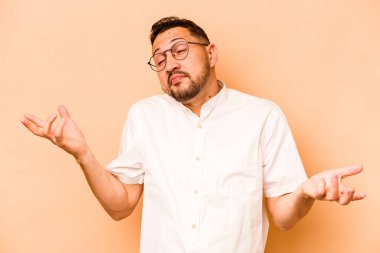 Latin kökenli genç bir adam, bej arka planda izole edilmiş sorgulama jestinde şüphe ediyor ve omuz silkiyor..