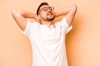 Latin kökenli genç adam bej arka planda izole edilmiş. Kendine güveniyor, elleri başının arkasında..