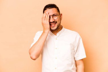 Latin kökenli genç adam bej arka planda izole edilmiş. Yüzünün yarısını avuç içiyle kaplayarak eğleniyor..