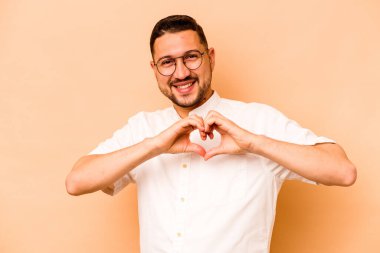 Latin kökenli genç adam bej arka planda izole edilmiş gülümsüyor ve elleriyle kalp şeklini gösteriyor..