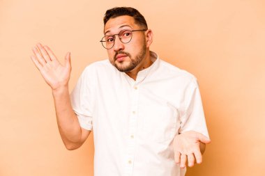 Latin kökenli genç adam bej arka planda izole edilmiş. Kopya alanını tutmak için kafası karışık ve omuz silkiyor..