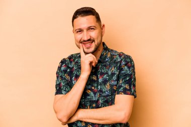 Latin kökenli genç adam bej arka planda izole edilmiş mutlu ve kendinden emin gülümsüyor, elleriyle çenesine dokunuyor..