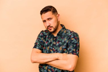 Latin kökenli genç adam bej arka planda izole edilmiş. Mutsuz, iğneleyici bir ifadeyle kameraya bakıyor..