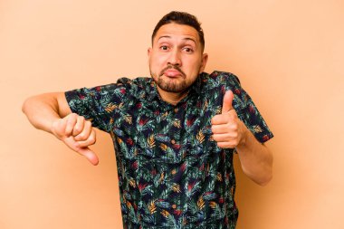Latin kökenli genç bir adam bej arka planda izole edilmiş. Baş parmağını kaldırıyor ve baş parmağıyla işaret ediyor. Zor seçim kavramı.