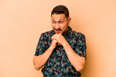 Latin kökenli genç adam bej arka planda izole edilmiş şans için dua ediyor, şaşırmış ve ağzını açıyor..