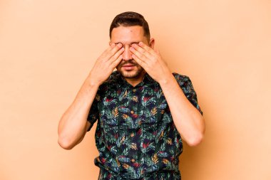 Latin kökenli genç bir adam bej arka planda izole edilmiş, elleriyle gözlerini kapatmaktan korkuyor..