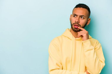 Genç İspanyol adam mavi arka planda izole edilmiş şüpheli ve şüpheci bir ifadeyle yana bakıyor..