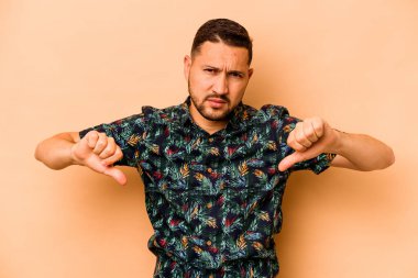 Latin kökenli genç adam bej arka planda izole edilmiş baş parmağını aşağı gösteren, hayal kırıklığı konsepti.