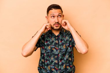 Latin kökenli genç adam bej arka planda izole edilmiş. Kulakları parmaklarıyla kapatıyor. Yüksek sesli bir ortam yüzünden stresli ve çaresiz..