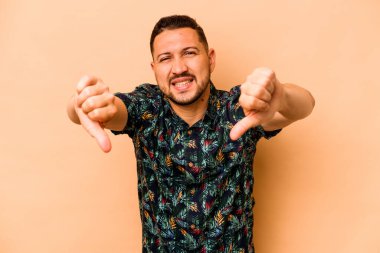 Latin kökenli genç adam bej arka planda izole edilmiş baş parmağını aşağı gösteriyor ve hoşlanmadığını ifade ediyor..