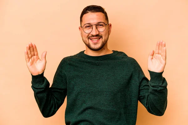 Latin kökenli genç adam bej arka planda izole edilmiş. Elinde küçük bir işaret parmağı olan, gülümseyen ve kendinden emin bir şey tutuyor..