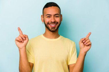 Mavi arka planda izole edilmiş genç İspanyol bir adam iki parmağı da boş bir alan gösteriyor..