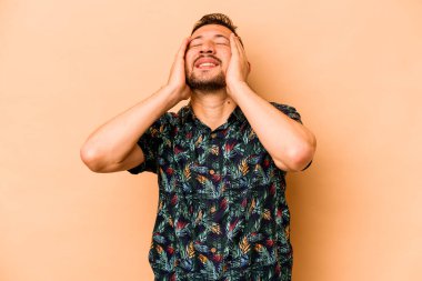 Latin kökenli genç bir adam bej arka planda yalnızken mutlu bir şekilde ellerini başının üstünde tutuyor. Mutluluk kavramı.