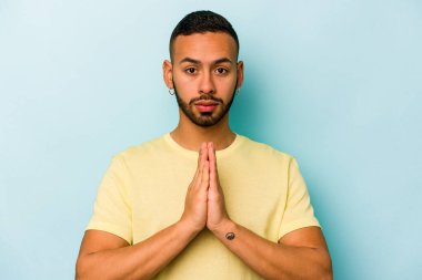 Genç İspanyol adam mavi arka planda izole edilmiş dua ediyor, bağlılık gösteriyor, dindar biri ilahi ilhamı arıyor..