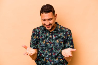 Latin kökenli genç adam bej arka planda izole edilmiş. Elinde avuç içi olan bir şey var ve kameraya sunuyor..