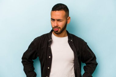 Mavi arka planda izole edilmiş, hoşnutsuzca kaşlarını çatmış, kollarını kavuşturmuş genç bir İspanyol..