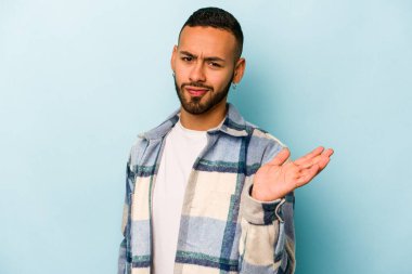 Latin kökenli genç bir adam, mavi arka planda izole edilmiş şüphe içinde ve sorgulama sırasında omuz silkiyor..