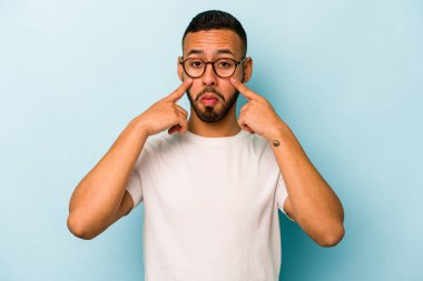 Genç İspanyol bir adam mavi arka planda izole edilmiş, ağlıyor, bir şeyden memnun değil, acı ve kafa karışıklığı konsepti..