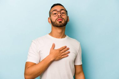 Genç İspanyol bir adam mavi arka planda izole edilmiş yemin ediyor, elini göğsüne koyuyor..