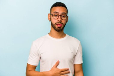 Mavi arka planda izole edilmiş genç İspanyol adam karnına dokunuyor, nazikçe gülümsüyor, yemek yiyor ve tatmin oluyor..