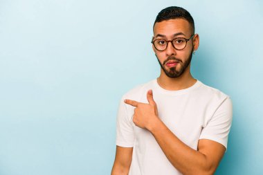Mavi arka planda izole edilmiş genç İspanyol adam gülümsüyor ve boş alanda bir şeyler gösteriyor..