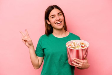 Pembe arka planda izole edilmiş patlamış mısır tutan beyaz bir kadın neşeli ve kaygısız parmaklarıyla barış sembolü gösteriyor..