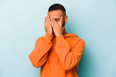 Mavi arka planda izole edilmiş genç İspanyol adam kameraya parmaklarıyla göz kırpıyor, yüzünü kapatmaktan utanıyor..