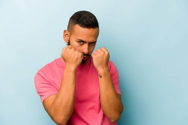 Latin kökenli genç bir adam mavi arka planda izole edilmiş. Yumruk atıyor, sinirleniyor, kavga ediyor..