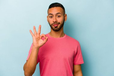 Mavi arka planda izole edilmiş genç İspanyol bir adam bir göz kırptı ve elleriyle iyi bir jest yaptı..