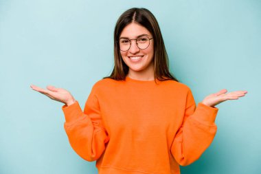 Mavi arka planda izole genç beyaz kadın kollarıyla ölçek yapıyor, mutlu ve kendinden emin hissediyor..