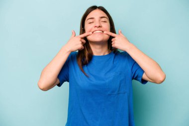 Mavi arka planda izole edilmiş genç beyaz kadın gülümsüyor, parmaklarını ağıza doğrultuyor..