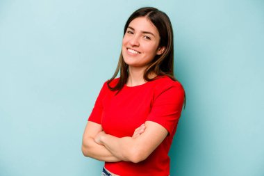 Mavi arka planda izole edilmiş genç beyaz kadın kendini güvende hissediyor, kararlılıkla kollarını kavuşturuyor..