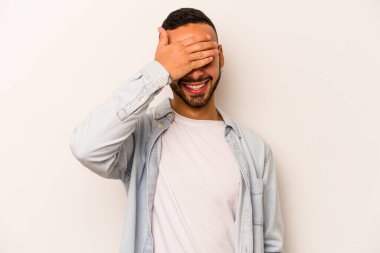 Beyaz arka planda izole edilmiş genç İspanyol adam elleriyle gözlerini kapatıyor, geniş bir gülümsemeyle bir sürpriz bekliyor..