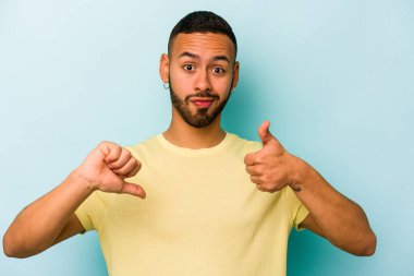 Latin kökenli genç adam mavi arka planda izole edilmiş, baş parmağını kaldırıyor, zor bir seçim konsepti.