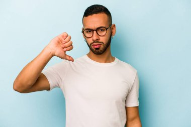 Mavi arka planda izole edilmiş genç İspanyol adam baş parmağını aşağı gösteriyor, hayal kırıklığı konsepti.