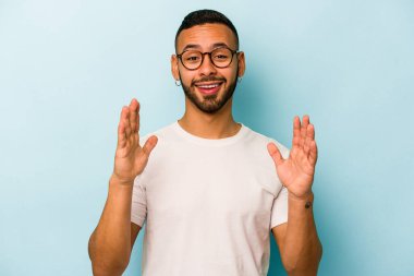 Mavi arka planda izole edilmiş genç İspanyol adam yüksek sesle gülüyor ve elini göğsüne koyuyor..