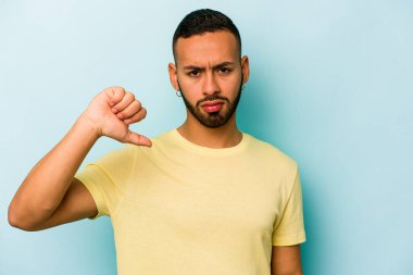 Mavi arka planda izole edilmiş genç İspanyol adam baş parmağını aşağı gösteriyor, hayal kırıklığı konsepti.