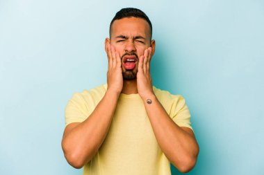 Genç İspanyol bir adam mavi arka planda izole edilmiş, ağlıyor, bir şeyden memnun değil, acı ve kafa karışıklığı konsepti..