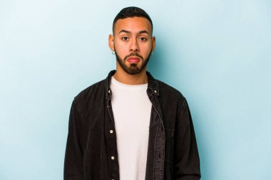 Mavi arka planda izole edilmiş genç bir İspanyol omuz silker ve gözleri açık kalır..