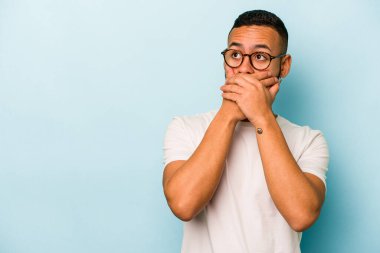 Genç İspanyol adam mavi arka planda izole edilmiş. Düşünceli bir şekilde bir fotokopi odasına bakıyor. Ağzını eliyle kapatıyor..