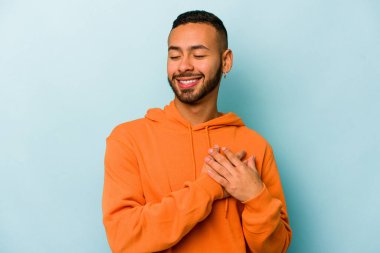 Mavi arka planda izole edilmiş genç İspanyol adam, avuçlarını göğsüne bastıran dostça bir ifadeye sahiptir. Aşk konsepti.