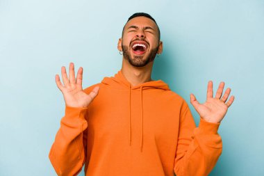 Genç İspanyol adam mavi arka planda izole edilmiş gökyüzüne haykırıyor, yukarı bakıyor, hayal kırıklığına uğramış..