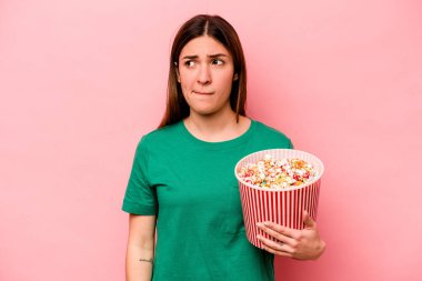 Pembe arka planda izole edilmiş patlamış mısır tutan genç beyaz kadın kafası karışmış, kuşkulu ve güvensiz hissediyor..