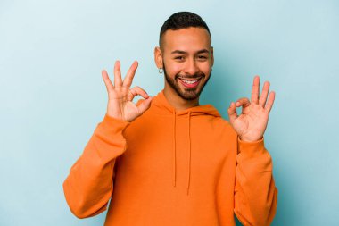 Genç İspanyol adam mavi arka planda izole edilmiş neşeli ve kendine güvenen iyi bir hareket sergiliyor..