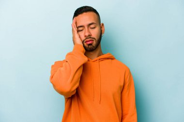 Genç İspanyol adam mavi arka planda izole edilmiş. Çok yorgun ve uykulu. Elini başının üstünde tutuyor..