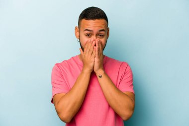 Mavi arka planda izole edilmiş genç İspanyol adam şok olmuş, ağzı ellerle kaplanmış, yeni bir şey keşfetmek için sabırsızlanıyor..