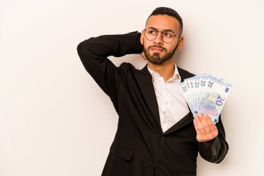 Genç İspanyol iş adamı, elinde banknotlar tutarken beyaz arka planda izole edilmiş kafasının arkasına dokunuyor, düşünüyor ve bir seçim yapıyor..